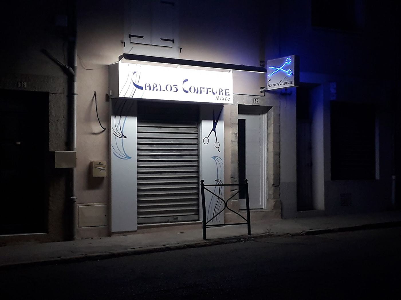 La façade du salon Carlos Coiffure de nuit, avec son enseigne lumineuse bleue en forme de ciseaux