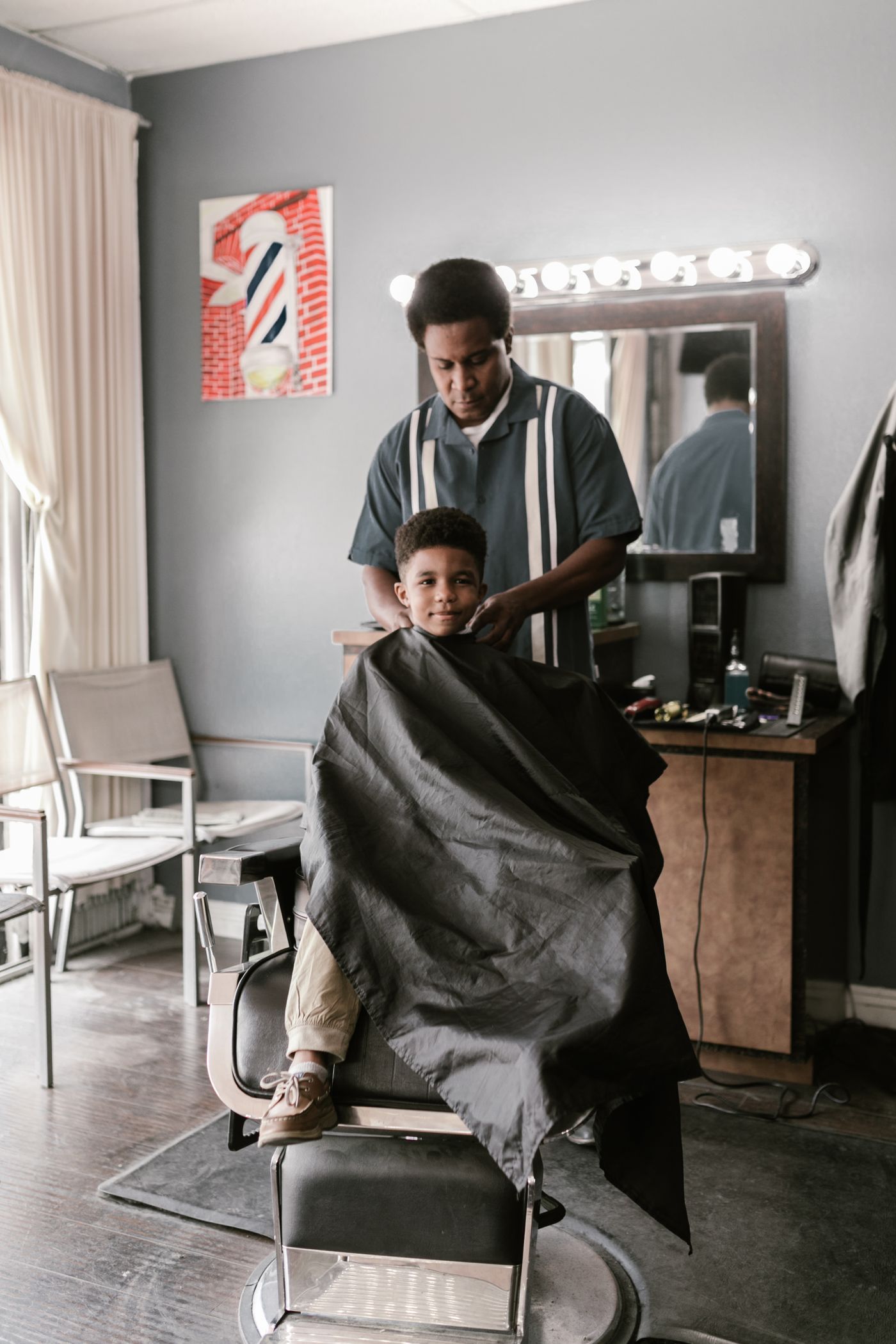 Un petit garçon installé dans le fauteuil sous une cape noire, pendant que le coiffeur prépare la coupe